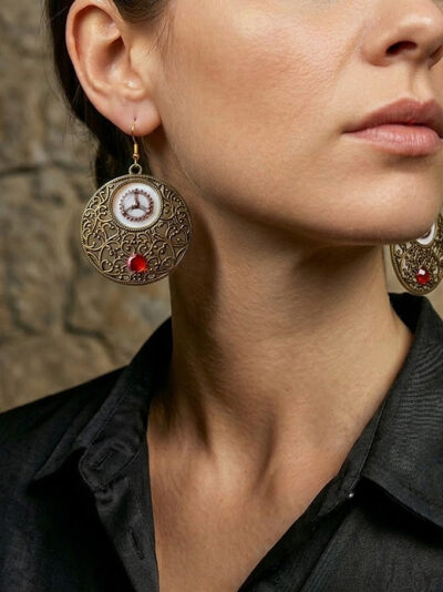 Boucles d’oreilles en Nacre surmontée d'un élément ouvragé et d'un engrenages en alliage bronze et de 2 beaux Grenats, avec crochets dorés portées par une femme brune aux cheveux attachés dont on ne voit que le bas du visage et qui porte une chemise noire