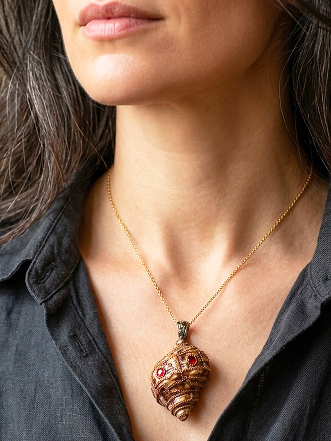 pendentif Coquillage marron foncé orné de perles dorées et de 5 Grenats avec une bélière gravée couleur bronze porté sur une chaine dorée par une femme brune aux cheveux longs dont on ne voit que le buste et qui porte une chemise noire ouverte