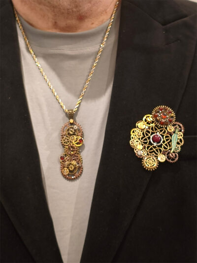 recto Broche steampunk composée d’engrenages coeurs et plume verte en alliage, ornée de 5 Grenats, 10 Agates rouges et marron et 1 Calcédoine  montée sur un axe porté par un homme sur tee shirt gris avec veste velours noire et on voit un collier engrenages également