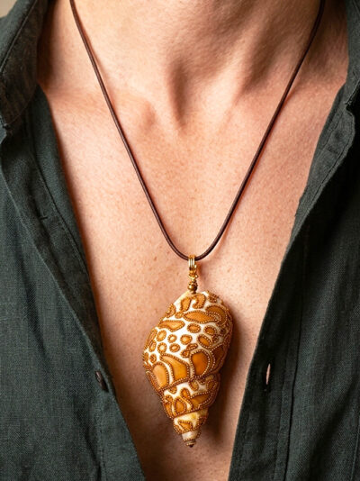 Pendentif Coquillage orné de perles dorées qui forment des tâches marron clair ressemblant à un jaguar avec bélière dorée porté sur un cordon de cuir foncé par un homme dont on ne voit que le buste et qui porte une chemise noire ouverte