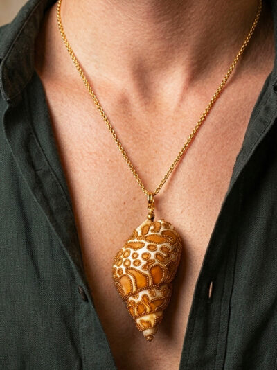 Pendentif Coquillage orné de perles dorées qui forment des tâches marron clair ressemblant à un jaguar avec bélière dorée porté sur une chaine mi longue dorée par un homme dont on ne voit que le buste et qui porte une chemise noire ouverte