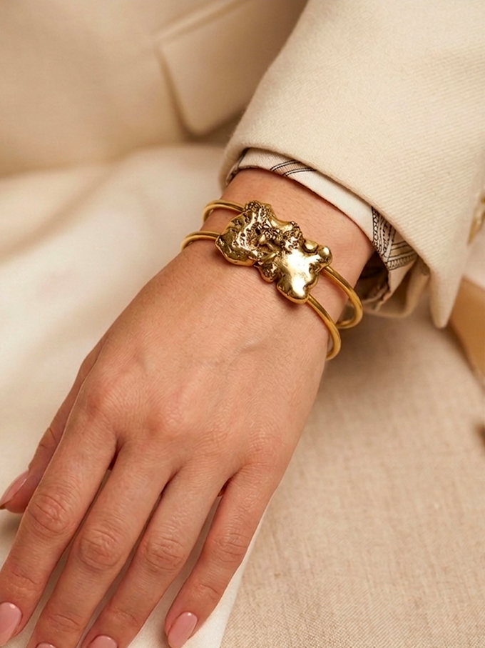 Bracelet manchette semi-ouvert en fil de laiton épais surmonté d'une pépite de laiton fondu de forme rectangulaire porté par une femme avec une veste beige dont on ne voit que la main, style chic