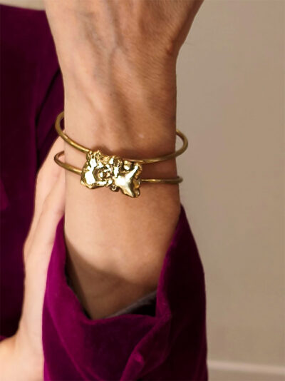 Bracelet manchette semi-ouvert en fil de laiton épais surmonté d'une pépite de laiton fondu de forme rectangulaire porté sur le poignet gauche par une femme portant une veste velours fuchsia dont on ne voit que l'avant bras