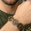 Bracelet en paracorde tressée avec des engrenages couleur bronze dessus et fil de laiton dessous porté par un homme brun barbu dont on ne voit que le poignet et le bas du visage, style steampunk
