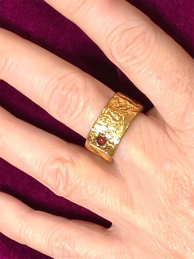 Bague semi-ouverte large en laiton réticulé avec un grenat incrusté sur le côté portée sur le majeur gauche d'une femme qui porte une veste velours fuchsia dont on ne voit que la main