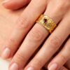Bague semi-ouverte large en laiton réticulé avec un grenat incrusté sur le côté portée par une femme dont on ne voit que la main
