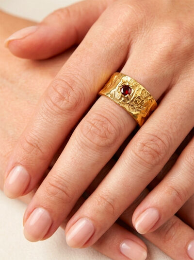 Bague semi-ouverte large en laiton réticulé avec un grenat incrusté sur le côté portée par une femme dont on ne voit que la main