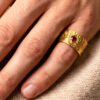 Bague semi-ouverte large en laiton réticulé avec un grenat incrusté sur le côté porté par un homme au petit doigt dont on ne voit que la main