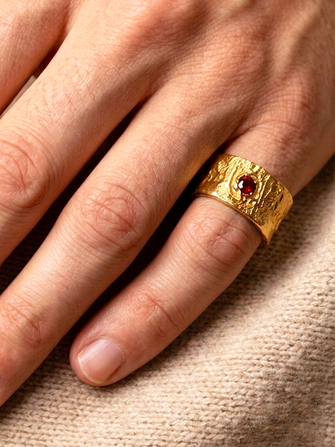 Bague semi-ouverte large en laiton réticulé avec un grenat incrusté sur le côté porté par un homme au petit doigt dont on ne voit que la main