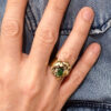 vue de face d'une bague fermée en laiton finition poli miroir avec pépite laiton fondue et perle de verre enserrée portée à l'annulaire droit d'une femme qui porte une chemise en jean dont on ne voit que la main posée sur la chemise