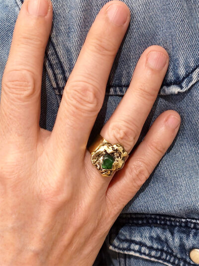 vue de face d'une bague fermée en laiton finition poli miroir avec pépite laiton fondue et perle de verre enserrée portée à l'annulaire droit d'une femme qui porte une chemise en jean dont on ne voit que la main posée sur la chemise