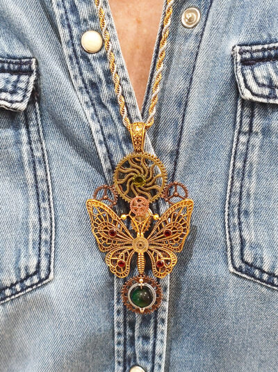 recto pendentif steampunk composé d'engrenages et d'un papillon en alliage orné d'un péridot vert et perle oeil de tigre vert montée sur axe et avec une bélière couleur bronze porté par une femme sur une chemise en jean