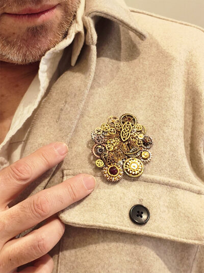 Recto d'une Broche steampunk composée d'engrenages et pièces en alliage ton dorés et cuivre avec des grenats et une perle calcédoine montée sur un axe portée par un homme à la barbe grise sur une veste en coton doux beige sur une chemise entrouverte