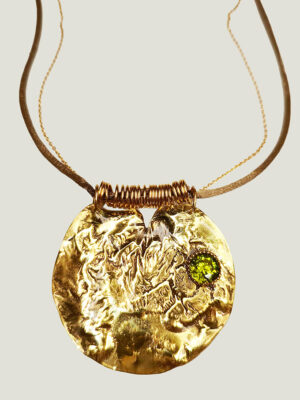 recto d'un pendentif de forme semi ronde en laiton réticulé avec un péridot sur le côté entouré de petites perles dorées, il est accroché à une chaîne dorée doublé d'un cordon en satin beige qui passe dans un rang d'anneaux dorés