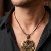Pendentif en alliage cuivre et laiton, orné d’engrenages et de 4 grenats avec bélière gravée bronze porté sur un cordon de cuir foncé court par un homme brun portant une chemise noire ouverte dont on ne voit que le buste et le bas du visage