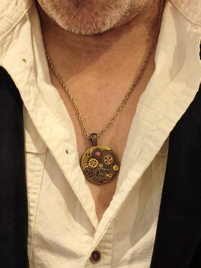 recto Pendentif en alliage cuivre et laiton, orné d’engrenages et de 4 grenats avec bélière gravée bronze porté par un homme dont on voit le bas du visage avec barbe grise avec chemise beige ouverte et veste noire velours