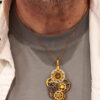 recto d'un pendentif steampunk composé d'engrenages en alliage orné d'un péridot vert et avec une bélière dorée porté par un homme sur un tee shirt gris avec chemise beige ouverte par dessus, on ne voit que le buste et bas du visage avec petite barbe grise de l'homme