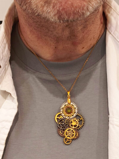 recto d'un pendentif steampunk composé d'engrenages en alliage orné d'un péridot vert et avec une bélière dorée porté par un homme sur un tee shirt gris avec chemise beige ouverte par dessus, on ne voit que le buste et bas du visage avec petite barbe grise de l'homme