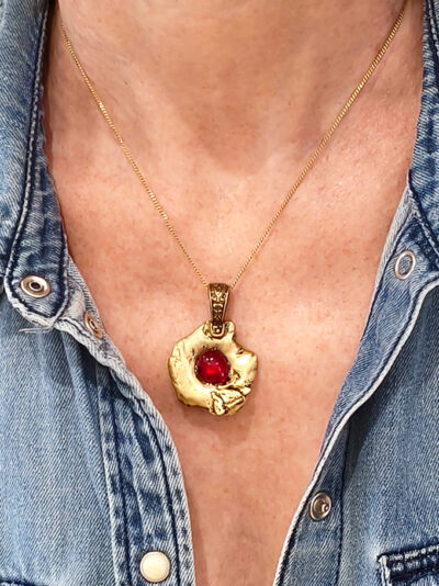 recto du pendentif en laiton fondu et formé en pépites avec un cabochon de verre rouge au centre. porté par une femme avec une chemise en jean bleue ouverte.