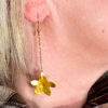 Boucles d'oreilles pendantes en forme de petites fleurs en laiton martelé attachées à des crochets dorés par une fine chaine dorée portées par une femme blonde sur l'oreille droite dont on ne voit que l'oreille et le bas du visage