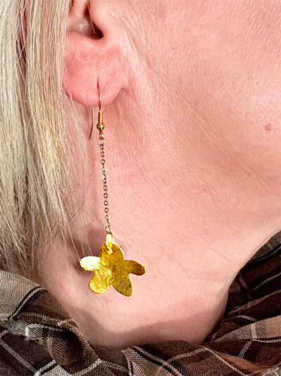 Boucles d'oreilles pendantes en forme de petites fleurs en laiton martelé attachées à des crochets dorés par une fine chaine dorée portées par une femme blonde sur l'oreille droite dont on ne voit que l'oreille et le bas du visage