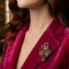 Orphélya P Broche engrenages plume en alliage avec des grenats et une perle calcédoine rose foncé porté par une femme brune au cheveu long dont on ne voit que le bas du visage sur le revers de sa veste en velours rose fuchsia