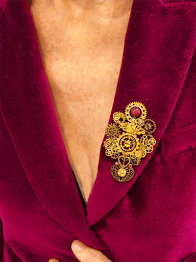 recto Broche engrenages plume en alliage avec des grenats et une perle calcédoine rose foncé porté par une femme portant une veste velours fuchsia dont on ne voit que le buste