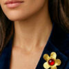Broche en laiton martelé en forme de grosse fleur avec un cabochon de verre rouge au centre entourée de petites perles dorées portée par une femme brune sur le revers gauche de sa veste en velours noir, style chic bohème