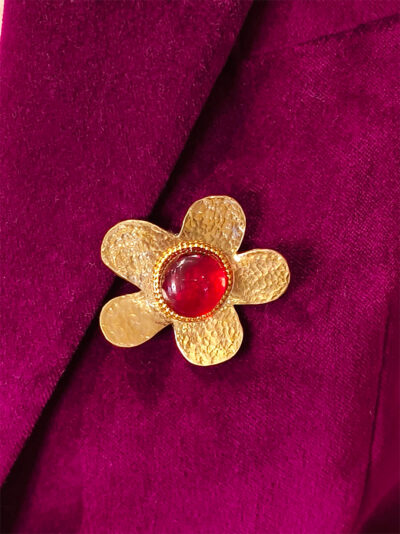 Recto d'une Broche en laiton martelé en forme de grosse fleur avec un cabochon de verre rouge au centre entourée de petites perles dorées portée sur une veste en velours rose fuchsia