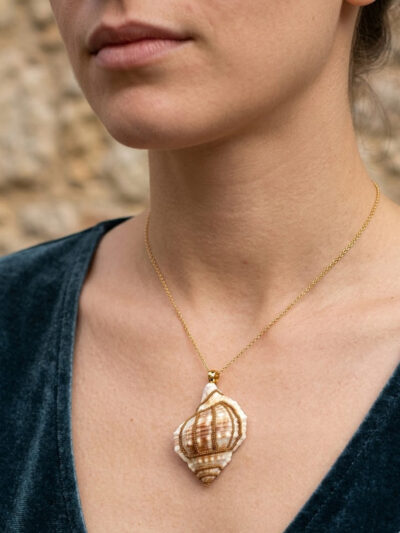 Pendentif Coquillage orné de perles dorées porté sur une chaine courte fine dorée par une femme brune aux cheveux attachés dont on ne voit que le bas du visage et le buste