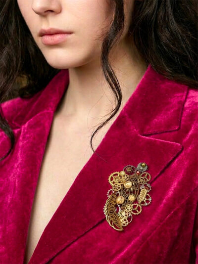 recto Broche engrenages et plume en alliage avec des grenats et une perle oeil de tigre verte portée par une femme brune au cheveu long dont on ne voit que le bas du visage sur le revers de sa veste en velours rose fuchsia