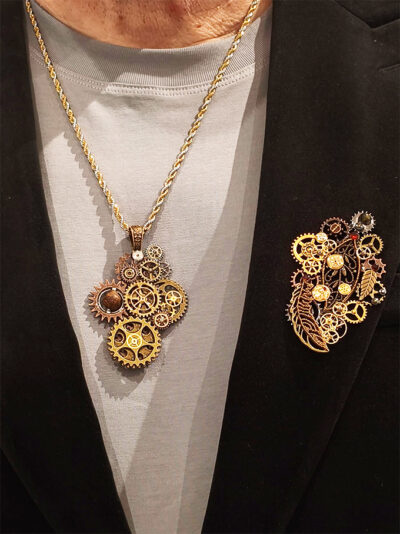 recto Broche steampunk engrenages et plume en alliage avec des grenats et une perle oeil de tigre vert porté par un homme sur un tee shirt gris sous une veste velours noir ouverte on voit également un collier steampunk