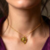 pendentif pépite laiton fondue ornée de petites perles dorées et d'une grenat avec anneau en laiton portée sur une chaine courte dorée par une femme brune aux cheveux attachés avec quelques mèches tombantes qui portent une chemise en lin violet
