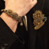Recto d'une broche steampunk faite avec engrenages et des pièces en laiton papillon et plume, soleil avec une perle verte montée sur un axe sur le côté et des grenats parsemés porté par un homme qui porte une veste noire et dont on ne voit que le bas du visage et la main qui porte un bracelet