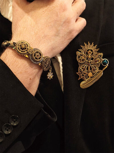 Recto d'une broche steampunk faite avec engrenages et des pièces en laiton papillon et plume, soleil avec une perle verte montée sur un axe sur le côté et des grenats parsemés porté par un homme qui porte une veste noire et dont on ne voit que le bas du visage et la main qui porte un bracelet