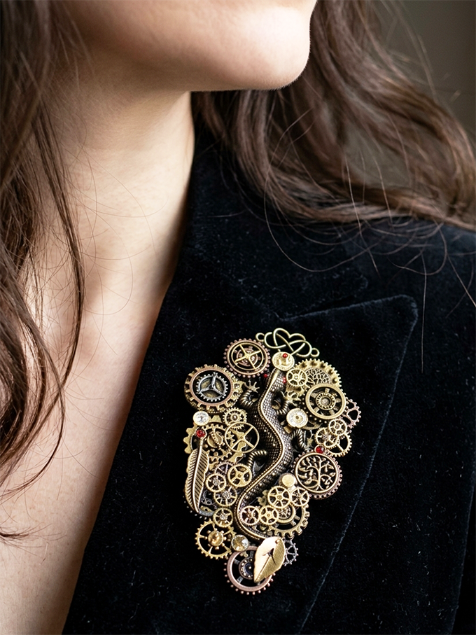 Broche engrenages et pièces en alliage ornée de 5 grenats et perles dorées portées par une femme ayant une veste velours noire