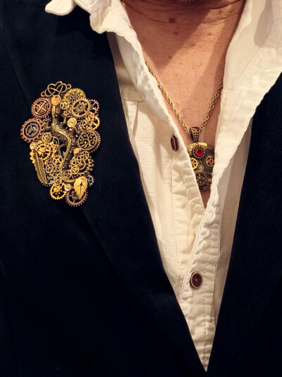 Broche steampunk avec engrenages et pièces en alliage, 5 grenats naturels et petites perles dorées portée par un homme sur le revers droit de sa veste en velours noir sur une chemise beige semi-ouverte sur un collier