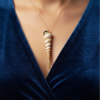 pendentif Coquillage très long et pointu beige clair orné de perles dorées et d'une obsidienne noire avec une bélière dorée porté sur une chaine dorée par une femme dont on ne voit que le buste et qui porte une robe en velours bleu foncé décolletée en V