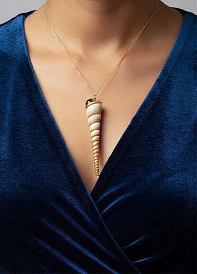 pendentif Coquillage très long et pointu beige clair orné de perles dorées et d'une obsidienne noire avec une bélière dorée porté sur une chaine dorée par une femme dont on ne voit que le buste et qui porte une robe en velours bleu foncé décolletée en V