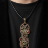 Pendentif composé d’engrenages alliage, orné de Grenats avec une bélière gravée de couleur bronze porté avec un cordon mi-long en cuir foncé par un homme dont on ne voit que le buste et qui porte un tee shirt noir