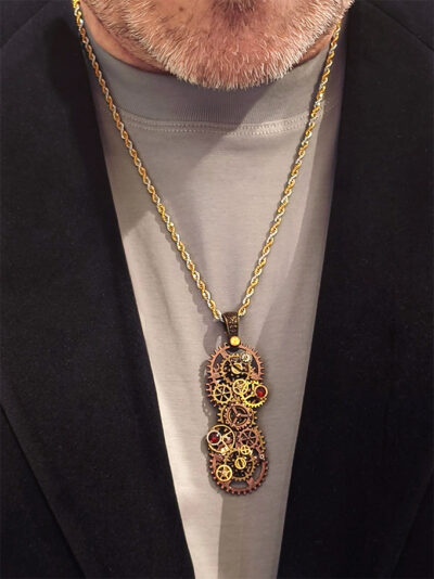 recto Pendentif long steampunk composé d’engrenages alliage, orné de Grenats avec une bélière gravée de couleur bronze porté par un homme dont on ne voit que le bas du visage avec barbe grise et buste sur tee shirt gris et veste noire velours