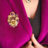 Broche engrenages coeur et plume en alliage avec des grenats et une perle calcédoine rose foncé portée par une femme dont on ne voit que le buste sur le revers d'une veste en velours rose fuchsia