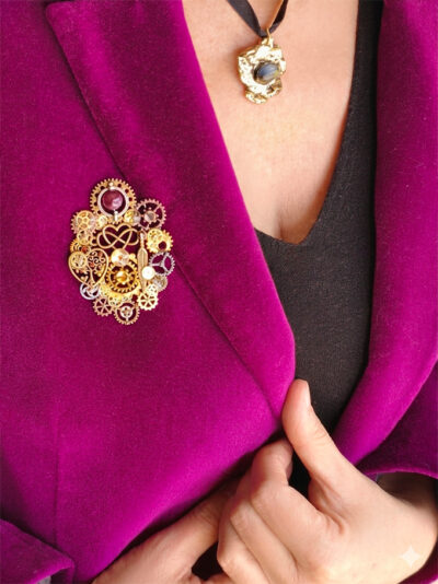 Broche engrenages coeur et plume en alliage avec des grenats et une perle calcédoine rose foncé portée par une femme dont on ne voit que le buste sur le revers d'une veste en velours rose fuchsia