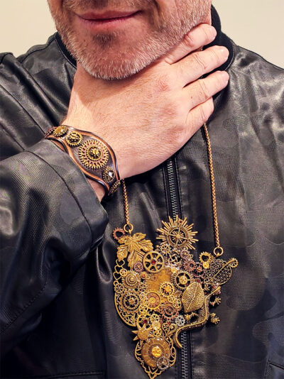 recto pendentif steampunk engrenages alliage salamandre couleur bronze ornés de 10 grenats attaché à 2 chaines doré porté par un homme aux cheveux et barbe grise dont on ne voit que le bas du visage et la petite barbe et qui porte un blouson en cuir noir fermé et au poignet droit devant son visage un bracelet cuir engrenages