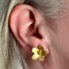 recto boucles d'oreilles clous en forme de fleur laiton doré avec 3 petites perles dorées portées sur l'oreille droite par une femme blonde dont on ne voit que l'oreille et une partie des cheveux