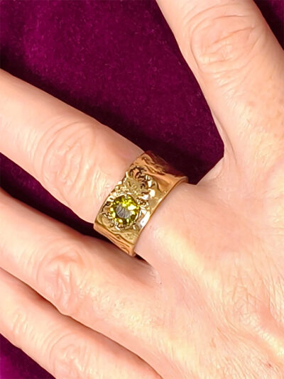 dessus de bague semi ouverte en laiton doré réticulé avec péridot vert portée par une femme au majeur gauche qui porte une veste velours fuchsia