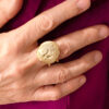vue dessus d'une bague en laiton doré avec dessus en pépites aspect brossé et nervuré et corps de bague en double fil de laiton portée au majeur de la main droite par une femme portant une veste fuchsia