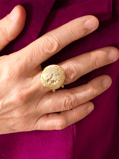 vue dessus d'une bague en laiton doré avec dessus en pépites aspect brossé et nervuré et corps de bague en double fil de laiton portée au majeur de la main droite par une femme portant une veste fuchsia