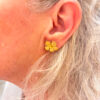 recto Boucles d'oreilles en forme de fleur en laiton brossé. Le centre de la fleur s'orne d'une petite perle dorée portée sur l'oreille gauche d'une femme avec cheveux mi-longs blonds/blancs