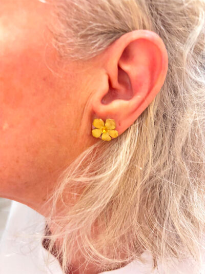recto Boucles d'oreilles en forme de fleur en laiton brossé. Le centre de la fleur s'orne d'une petite perle dorée portée sur l'oreille gauche d'une femme avec cheveux mi-longs blonds/blancs